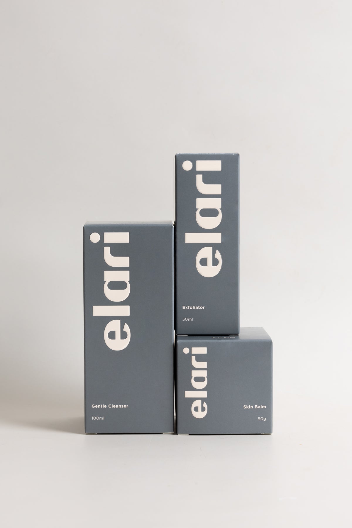 Three dark gray Elari product boxes stacked together on a light gray studio background. The boxes are branded 'elari' in large white vertical lettering, with smaller white text below indicating 'Gentle Cleanser', 'Exfoliator', and 'Skin Balm'.