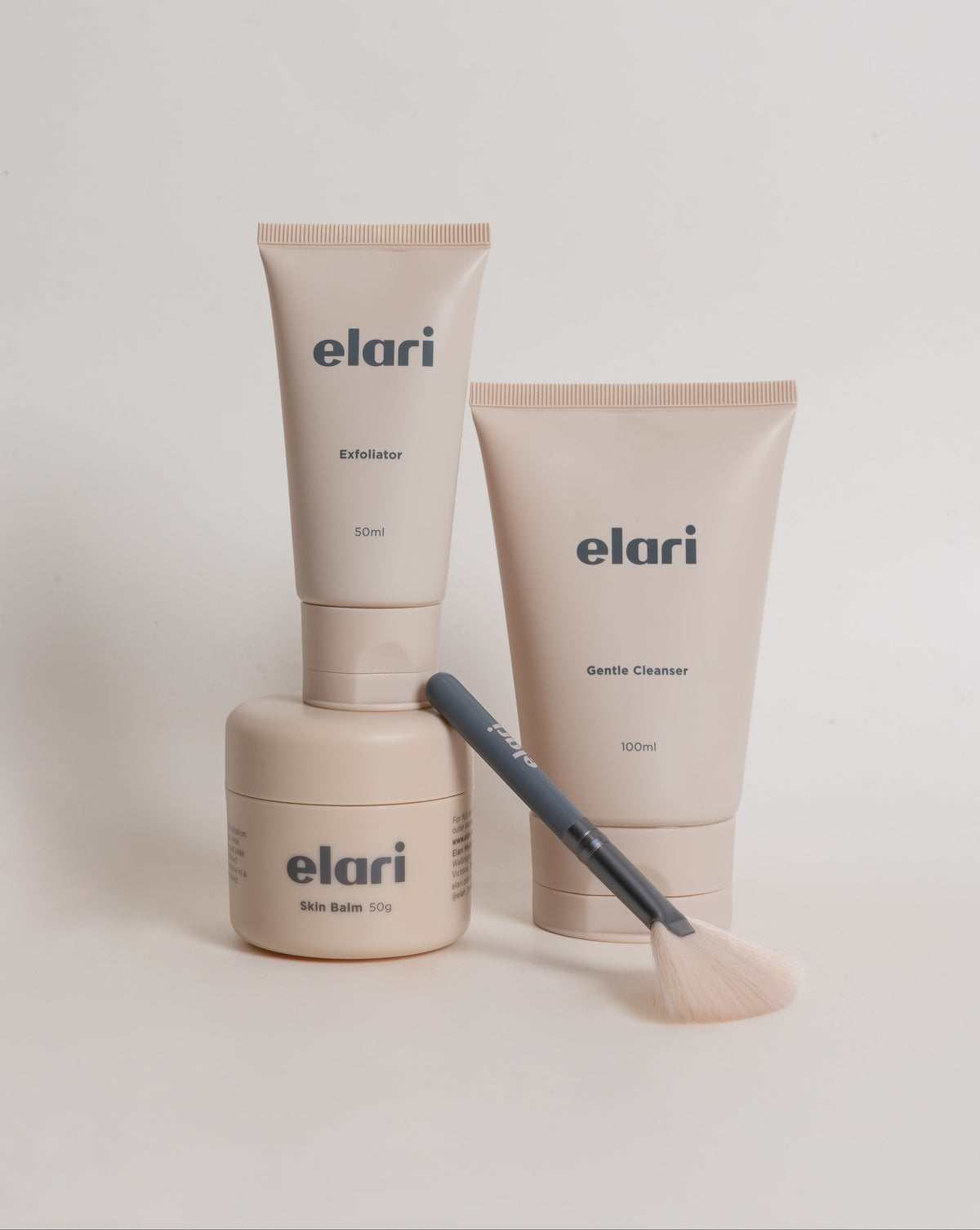 Three  skincare products from elari are arranged on a light gray background: an Exfoliator tube, a Cleanser tube, and a Skin Balm jar, with an exfoliator brush resting across the jar.